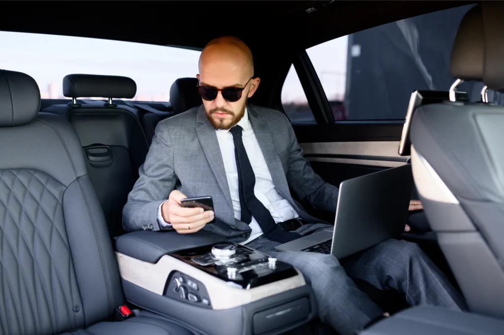 Businessmen watching phone while sitting in Black Car with laptop on his lap.